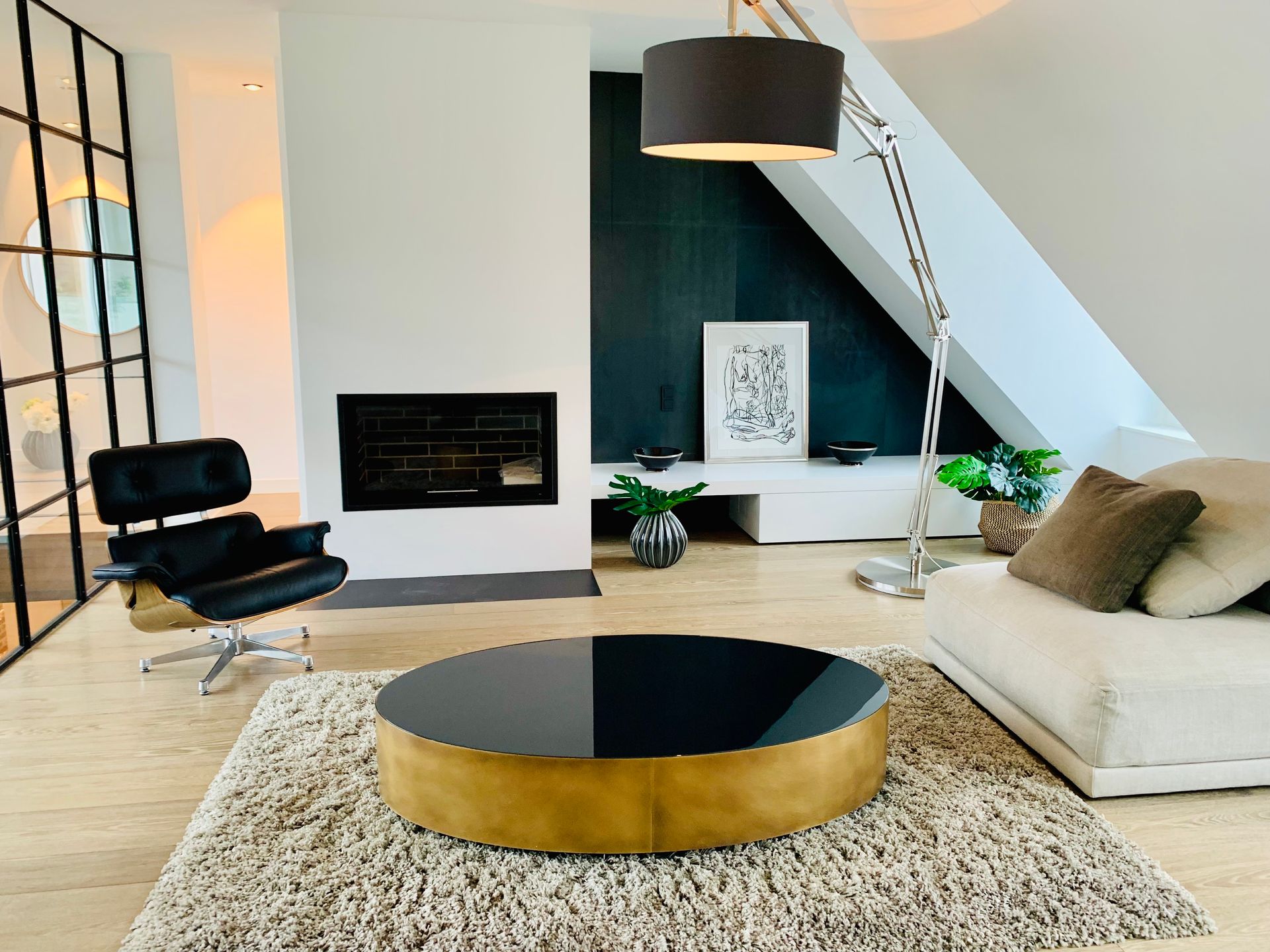 Modern living room with black fireplace, tan sofa, and round gold coffee table beneath a sloped ceiling