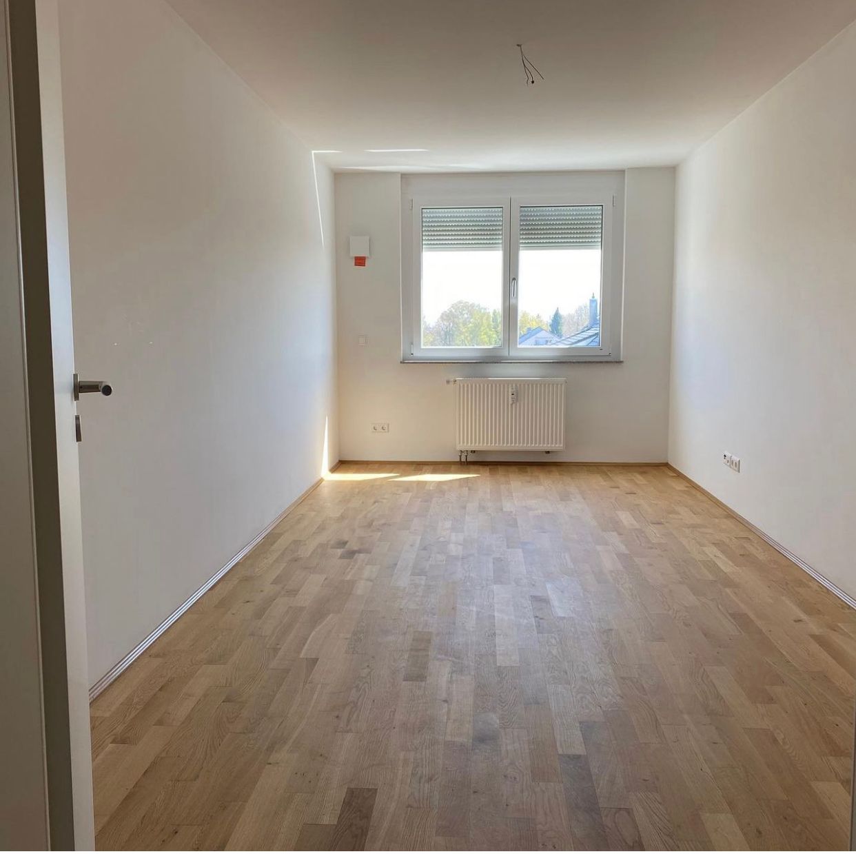 Empty rectangular room with light wood floor, white walls, and a window above a radiator.