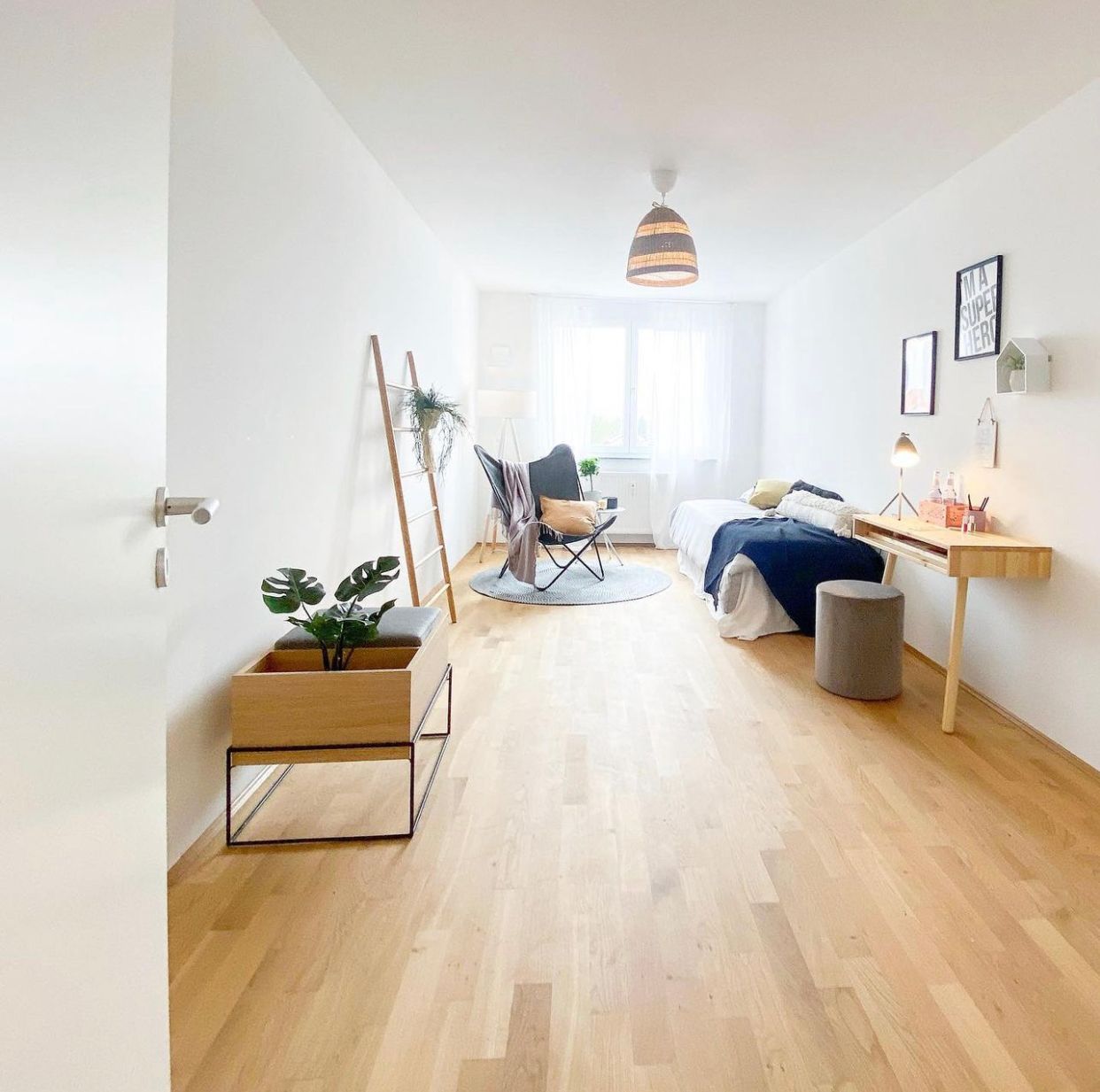 Bright minimalist bedroom with wood floor, blue bed, desk, plant, chair, and hanging lamp.