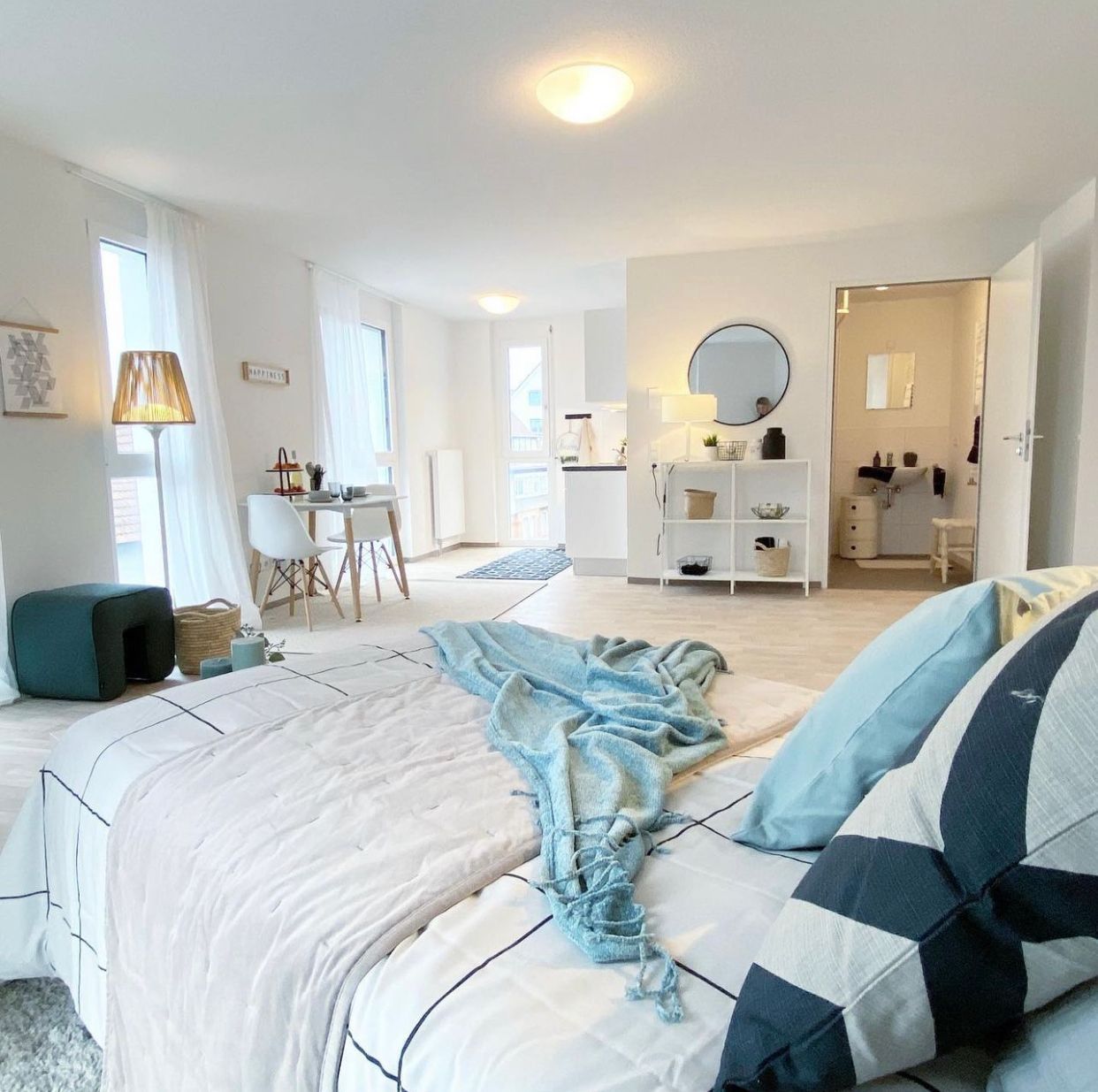 Bright bedroom with teal accents, a bed, vanity, lamp, and small dining area in the background