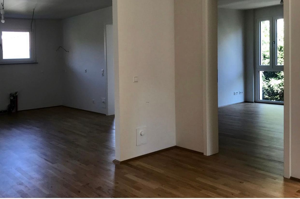 Empty living room with wood floors, white walls, and a doorway to a brighter adjacent room