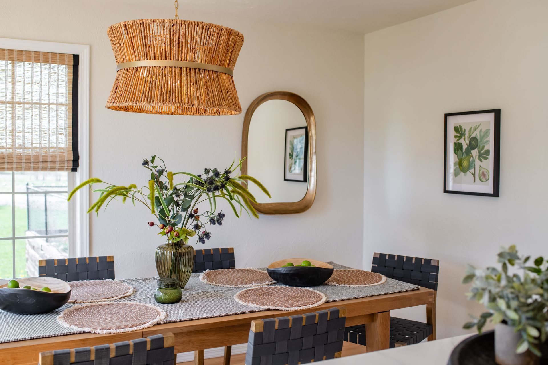 Bright dining room with wooden table, woven pendant lamp, round mirror, and plants by large windows
