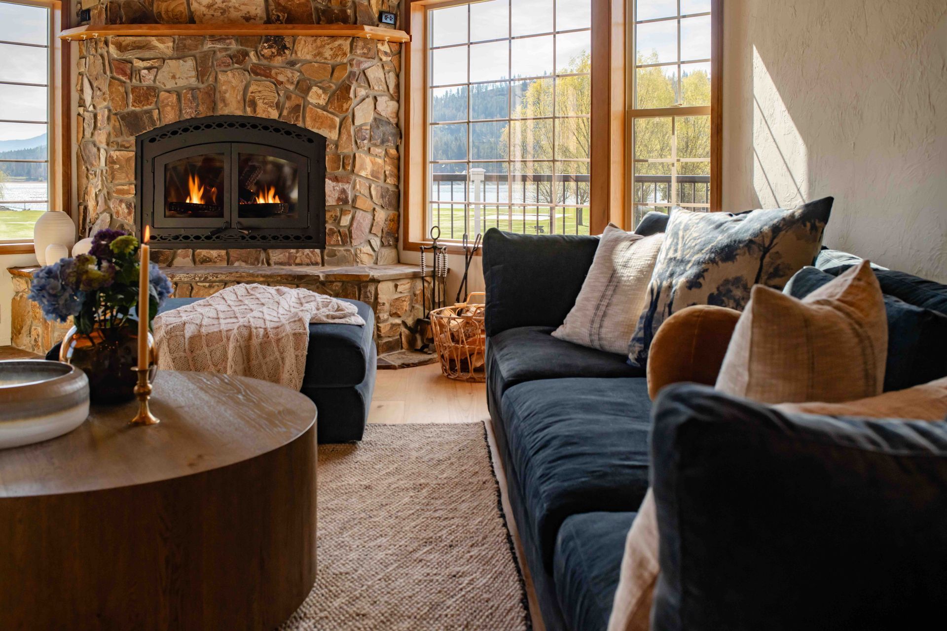 Cozy living room with stone fireplace, navy sofa, round wooden table, and large sunlit windows