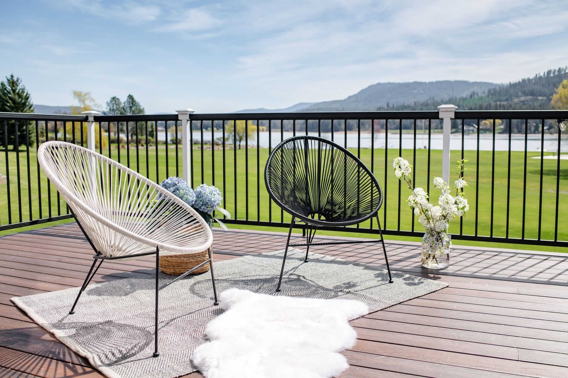 Two patio chairs on a deck by a black railing, overlooking water, hills, and mountains.