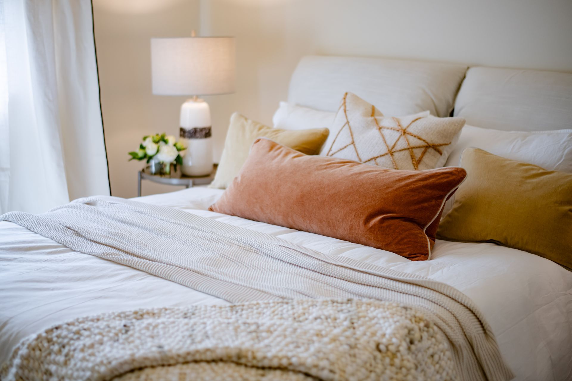 Neatly made bed with white bedding, beige pillows, and a bedside lamp in a bright bedroom
