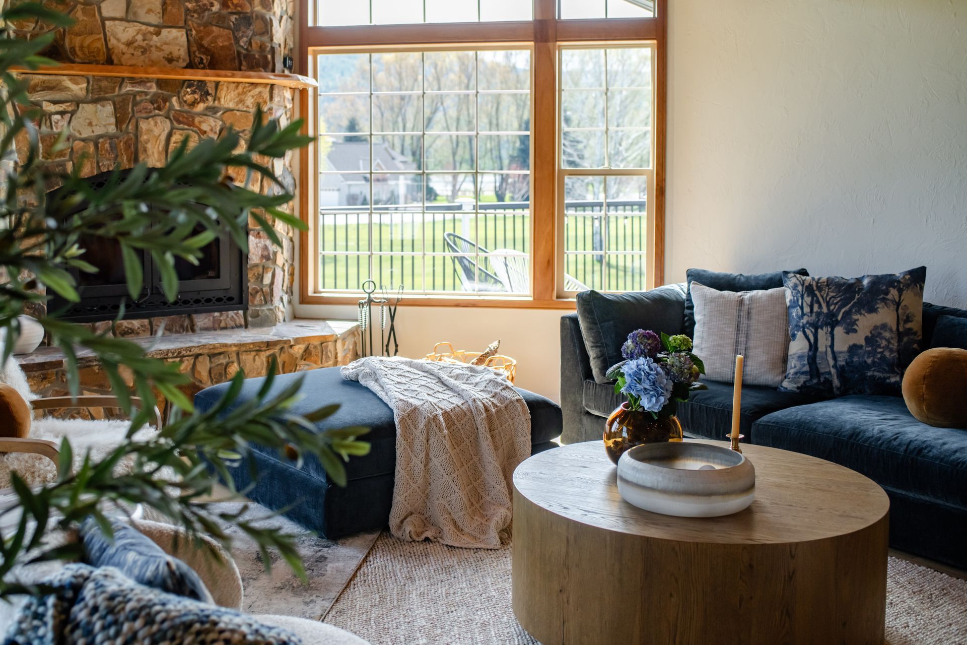 Cozy living room with stone fireplace, blue sofa, round wood table, and large window with outdoor view