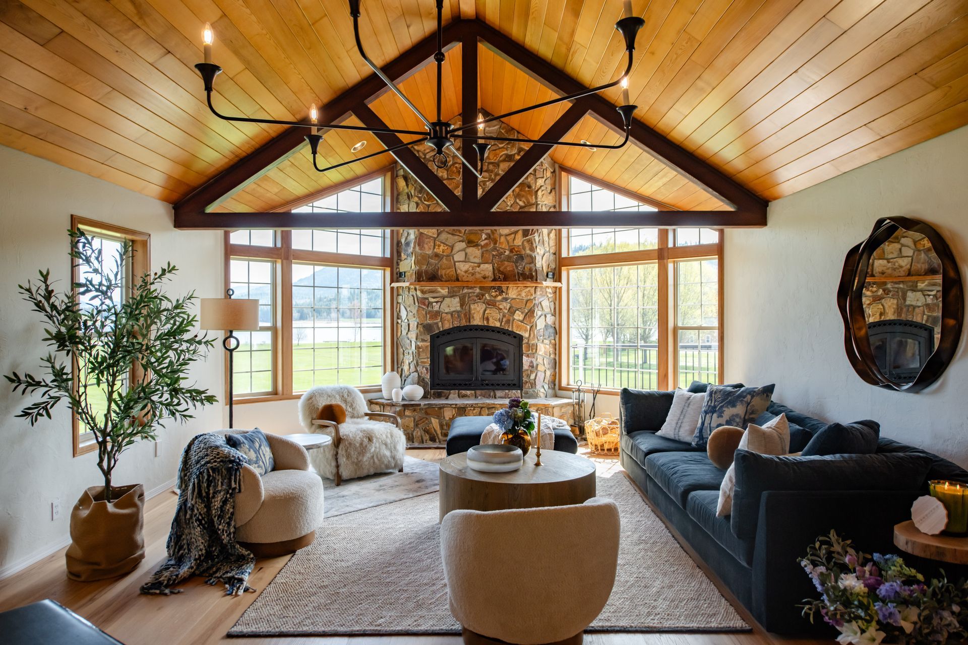 Cozy living room with vaulted wood ceiling, stone fireplace, large windows, and dark sectional sofa