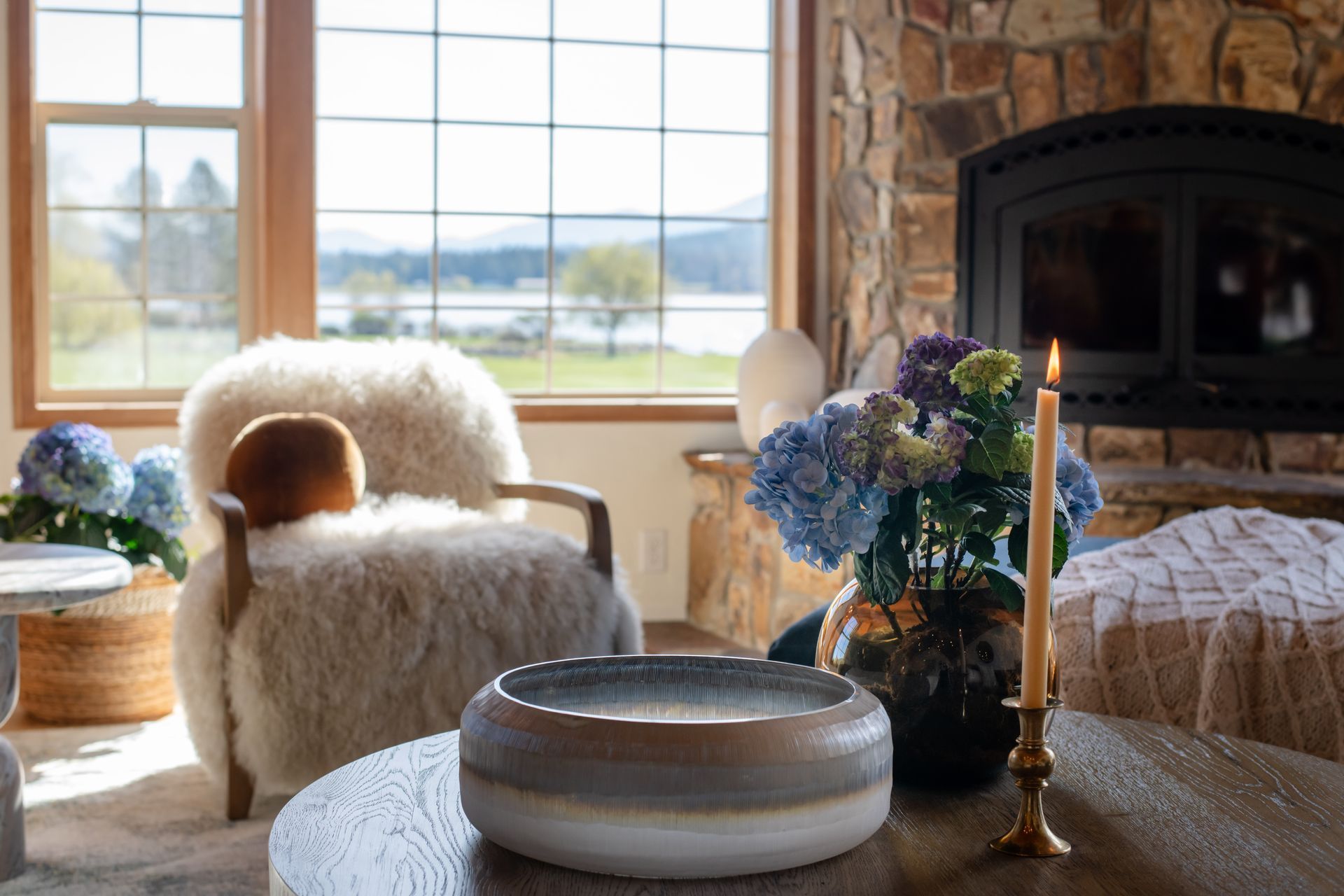 Cozy living room with fur chairs, stone fireplace, blue hydrangeas, and soft natural light from large windows.