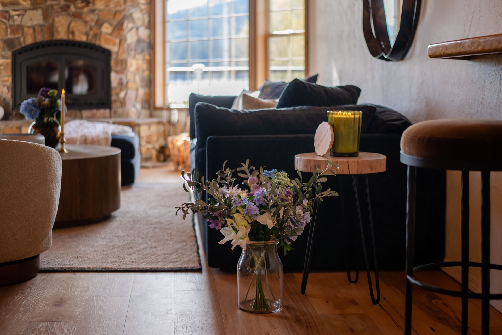 Cozy living room with stone fireplace, dark sofa, window light, and a flower arrangement on a small table.