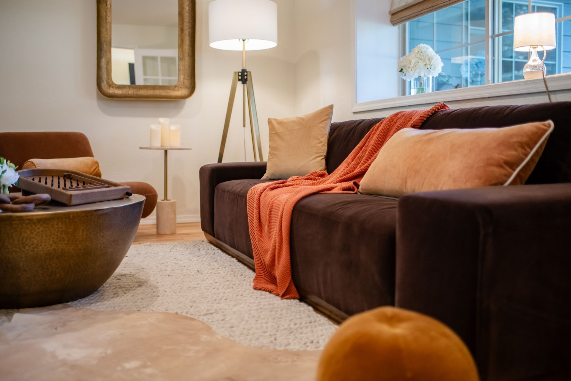 Cozy living room with brown sofa, orange throw, floor lamp, mirror, and window light
