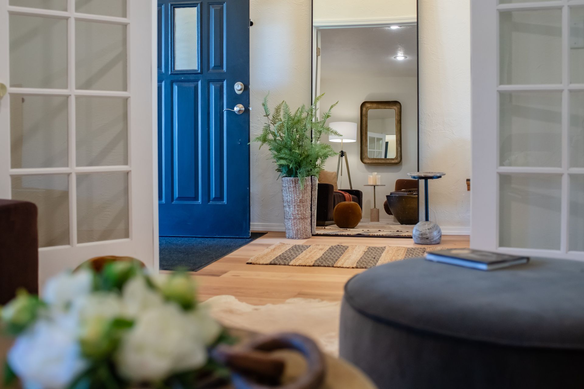 Bright living room with a blue front door, white shelving, flowers, and a mirror by the entryway.