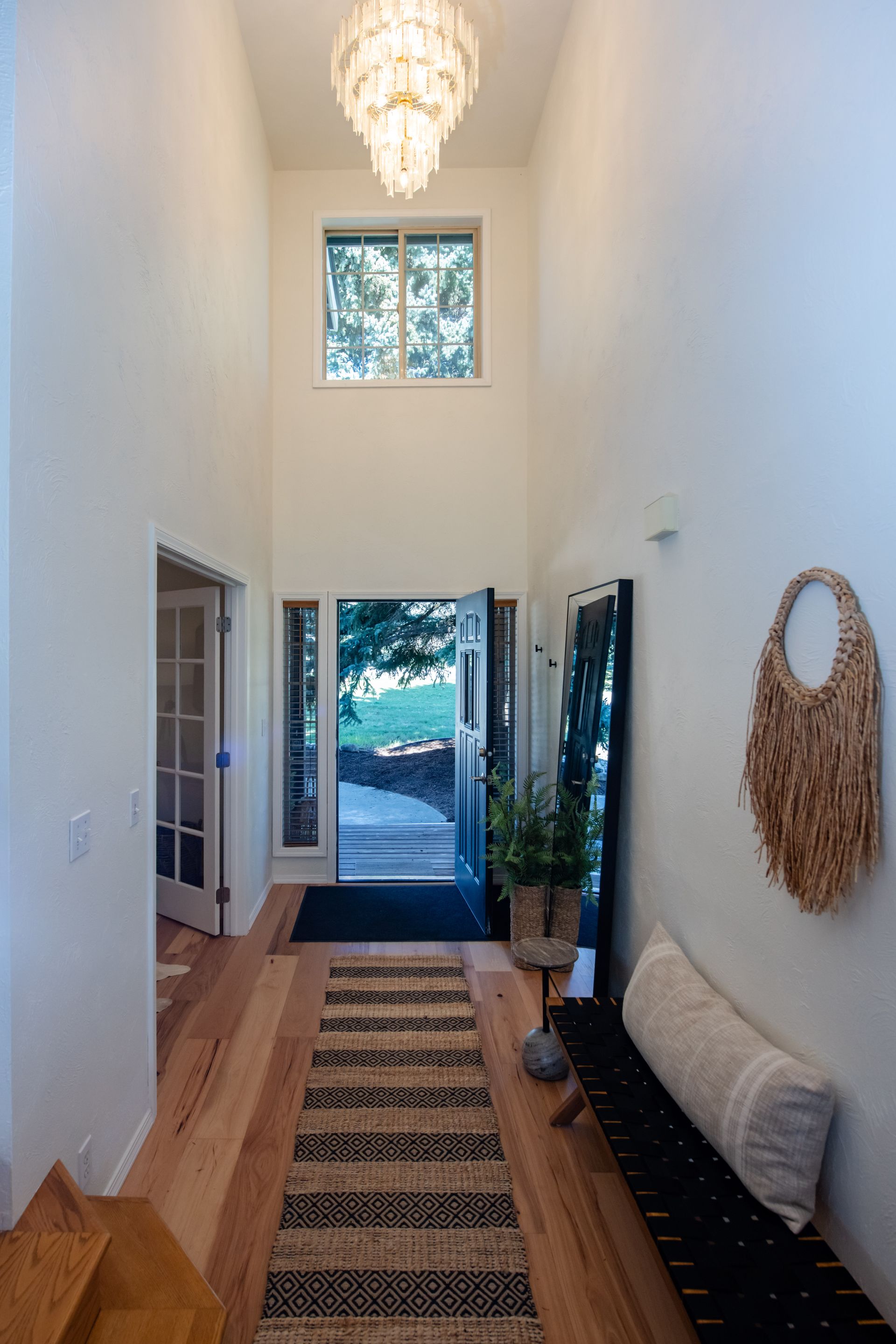 Narrow entry hall with tall white walls, patterned runner, chandelier, and open front door to daylight