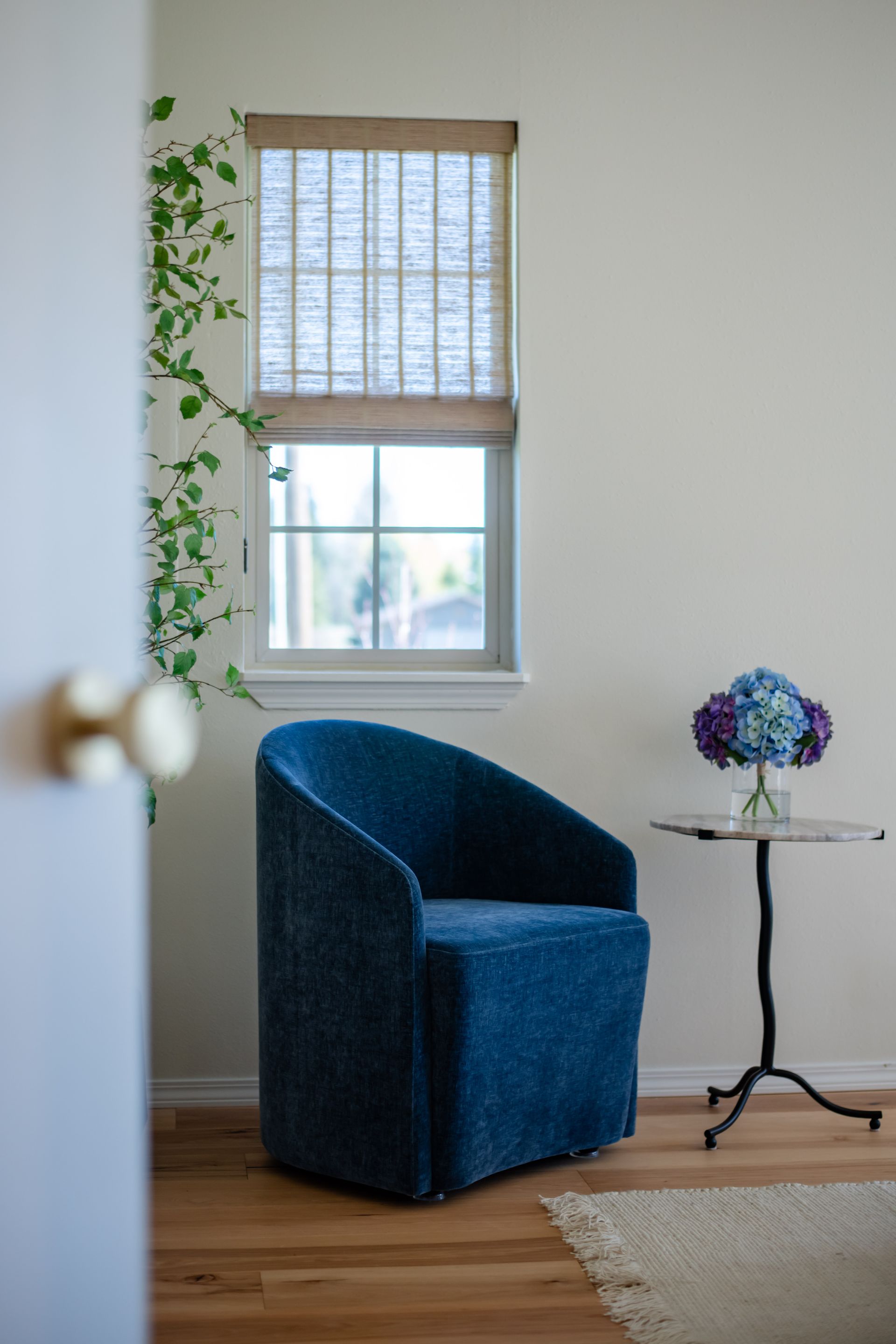 Blue upholstered chair by a window with bamboo blinds, next to a small glass table and purple flowers.