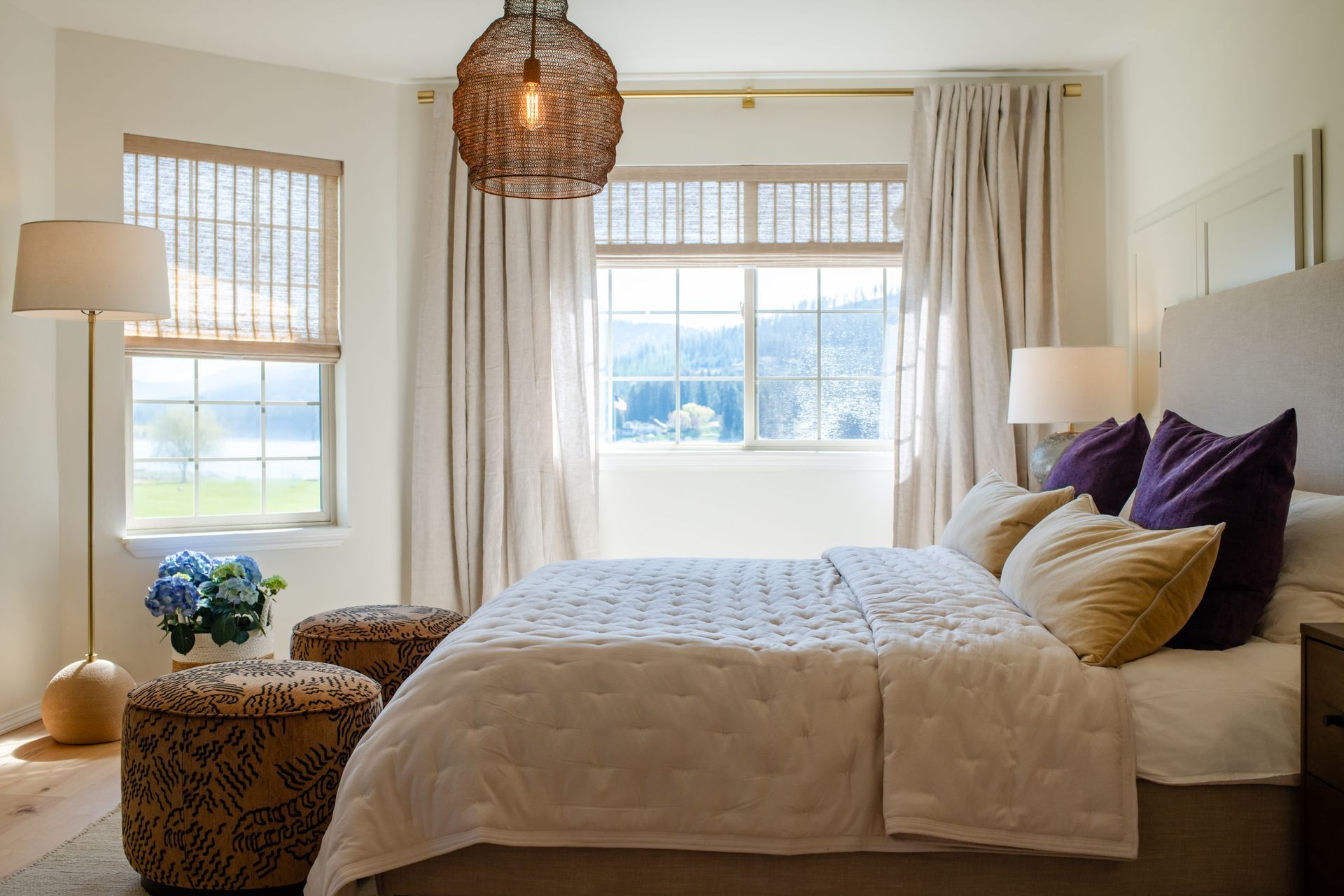 Bright bedroom with a white bed, purple pillows, wicker pendant light, and large sunlit windows