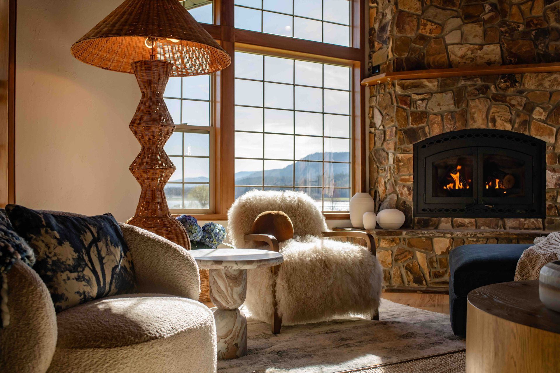 Cozy rustic living room with stone fireplace, fur chairs, large window, and warm lamp glow