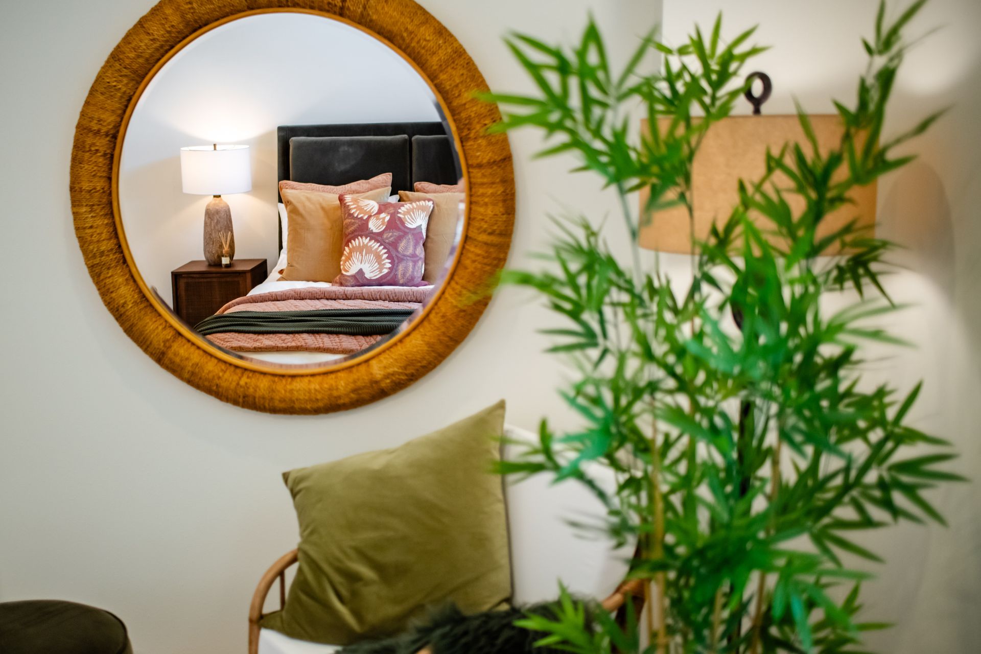 Round mirror reflecting a bed with pink pillows beside a green plant in a bright room