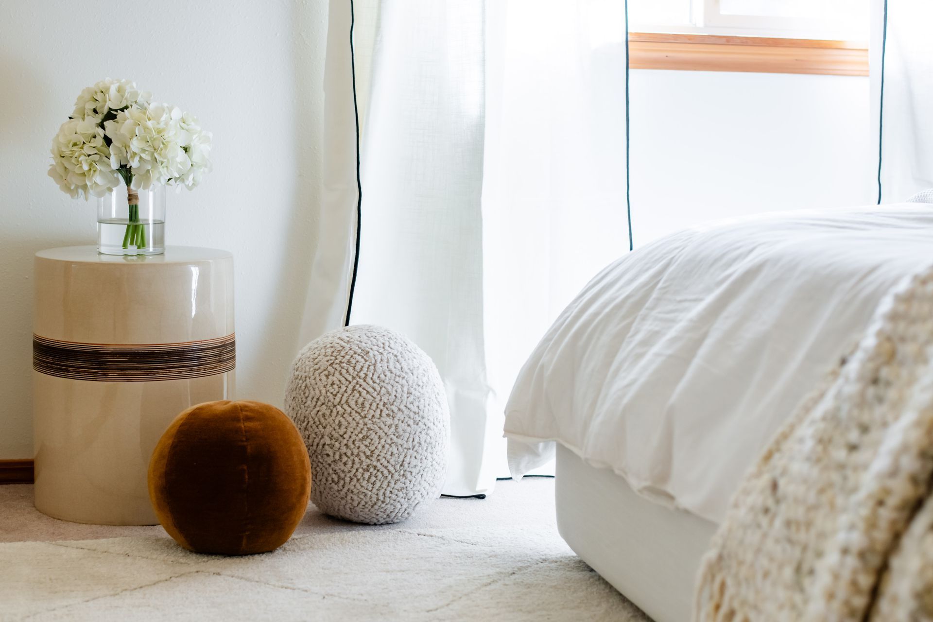 Cozy white bedroom corner with round stools, a flower vase, sheer curtains, and a bed with textured blanket
