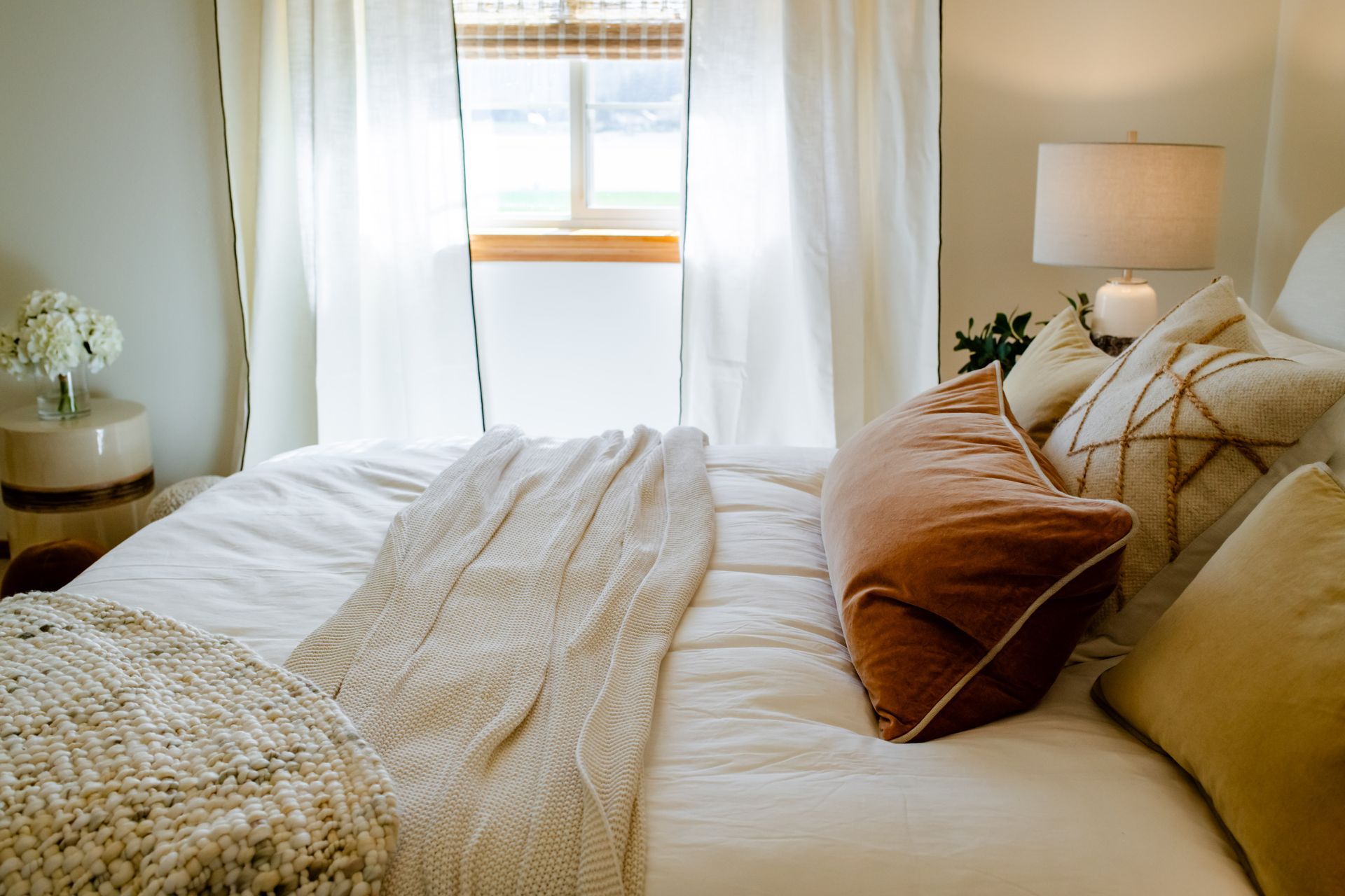 Bright cozy bedroom with a white bed, beige pillows, and sheer curtains over a sunlit window