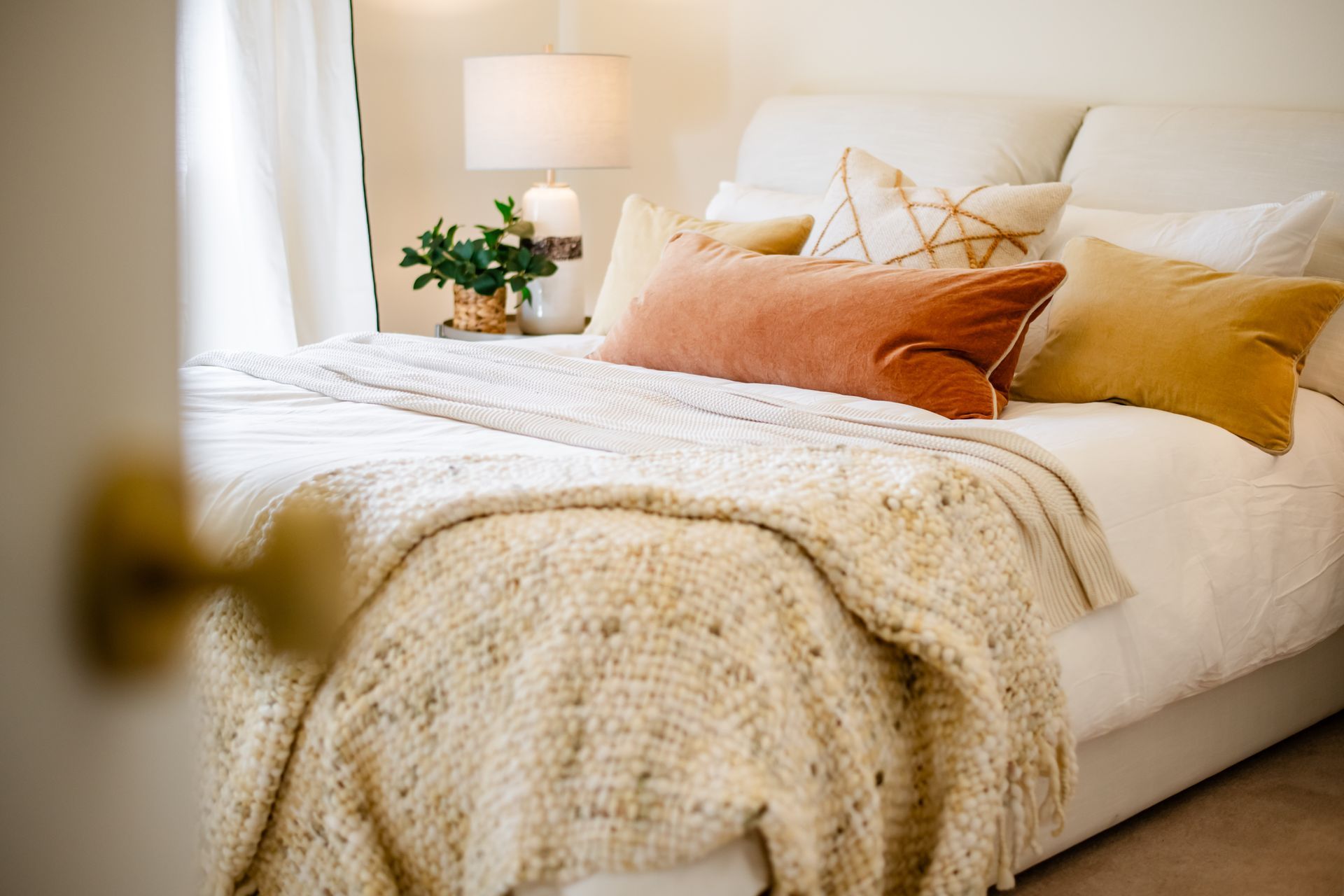 Cozy bed with white bedding, orange and beige pillows, soft throw blanket, and a bedside plant near a window