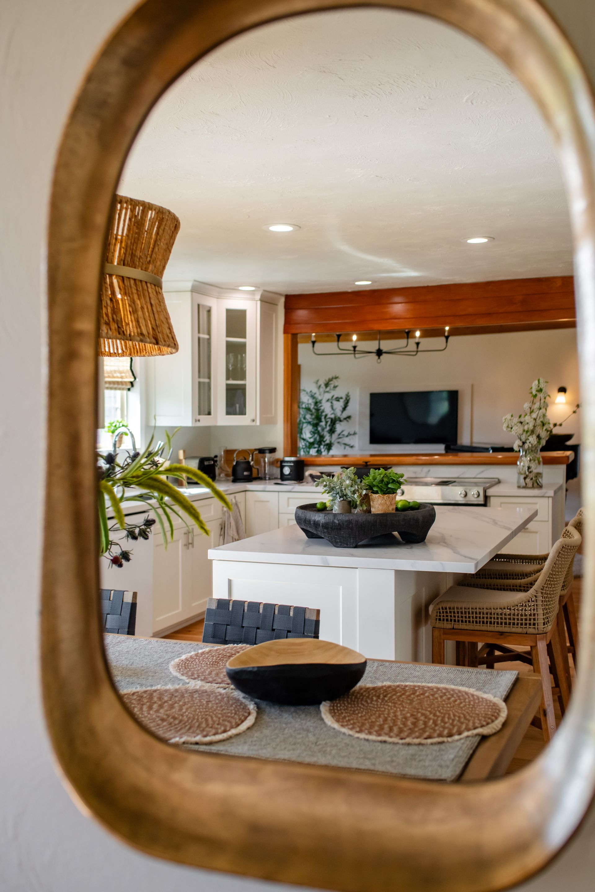 Dining room viewed through an oval mirror, with a white table, woven chairs, and a kitchen beyond.