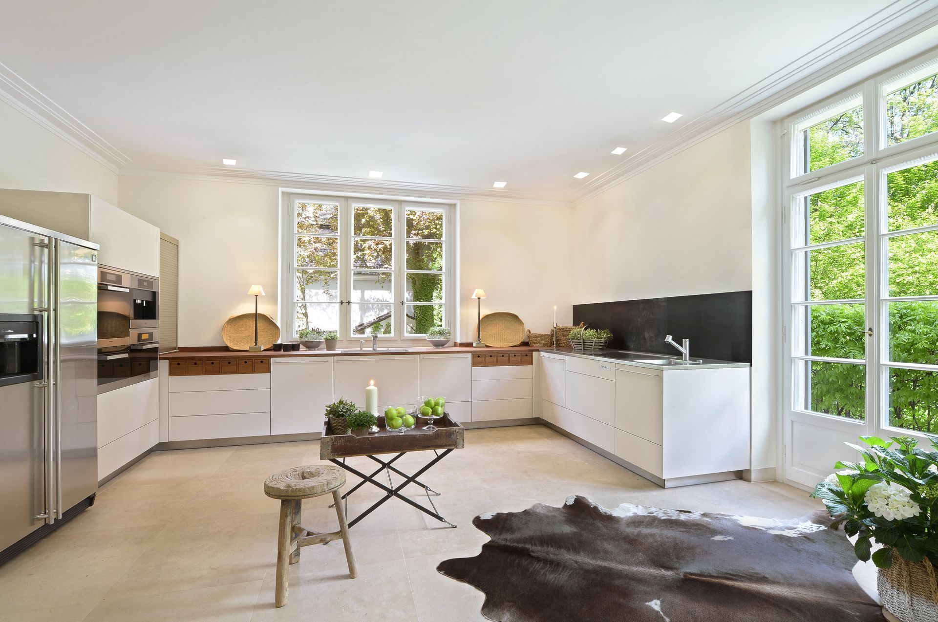Bright modern kitchen with white cabinets, stainless fridge, wood counters, and large windows overlooking greenery