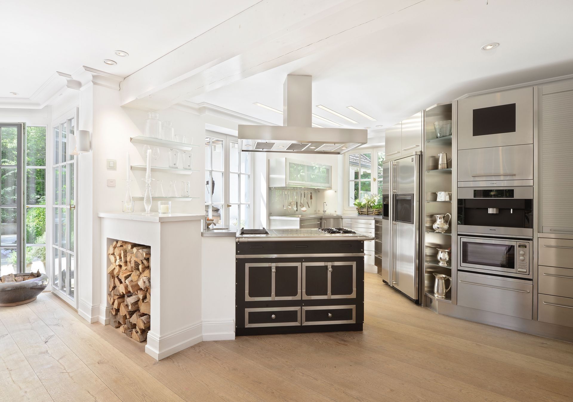 Bright modern kitchen with white walls, island, built-in ovens, and large windows showing greenery