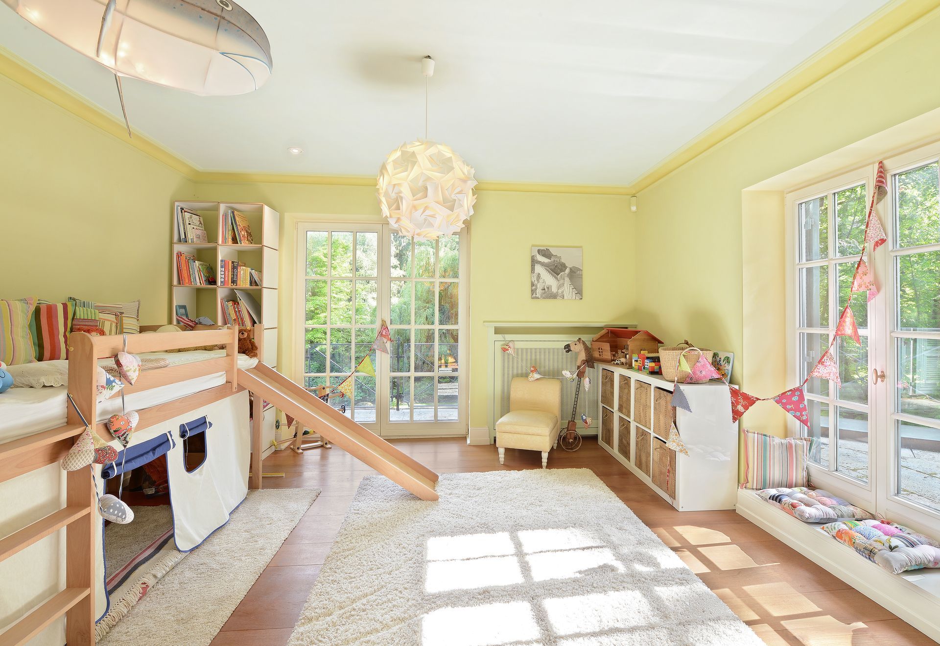 Bright child’s bedroom with pale yellow walls, loft bed, desk, and large windows letting in sunlight.