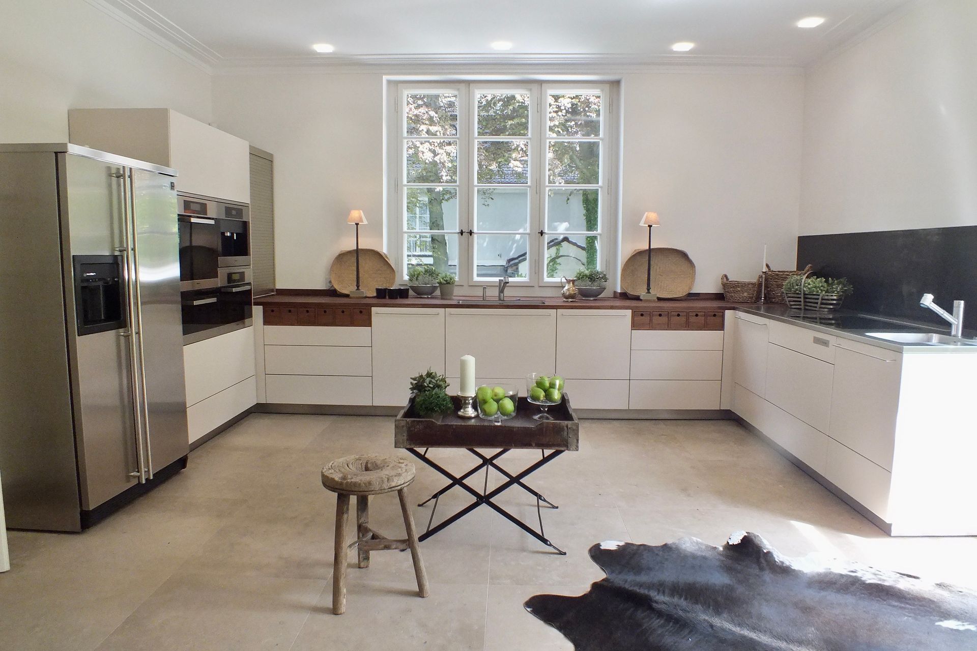 Bright white modern kitchen with stainless steel appliances, large window, and a small table with plants.