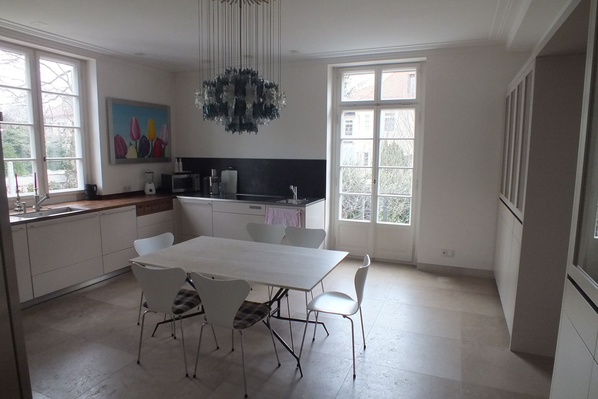Bright dining room with white table, chairs, large windows, chandelier, and compact kitchen area
