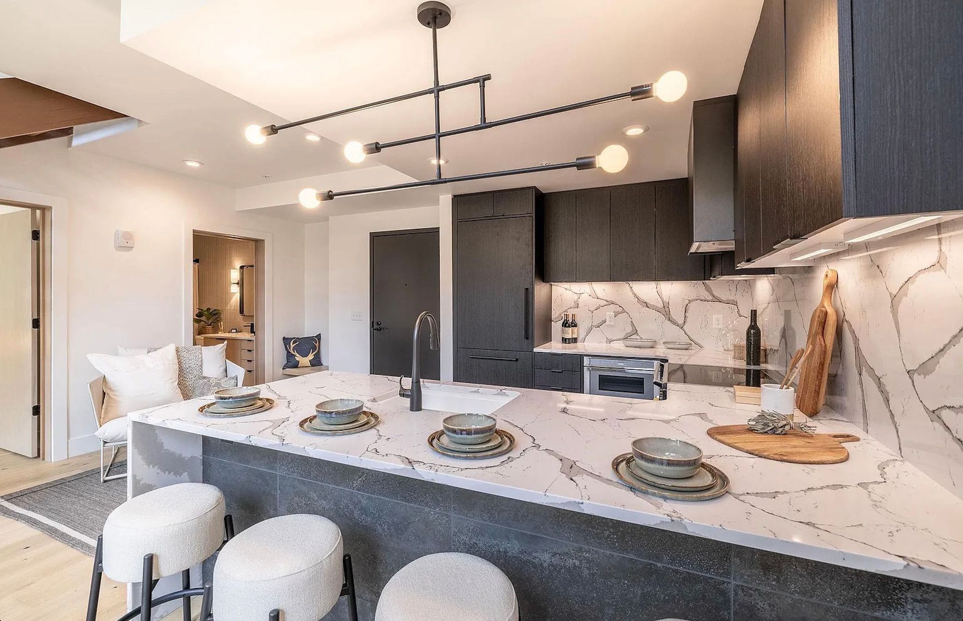 Modern open-plan kitchen with a large island, white stools, and dark cabinets under pendant lights
