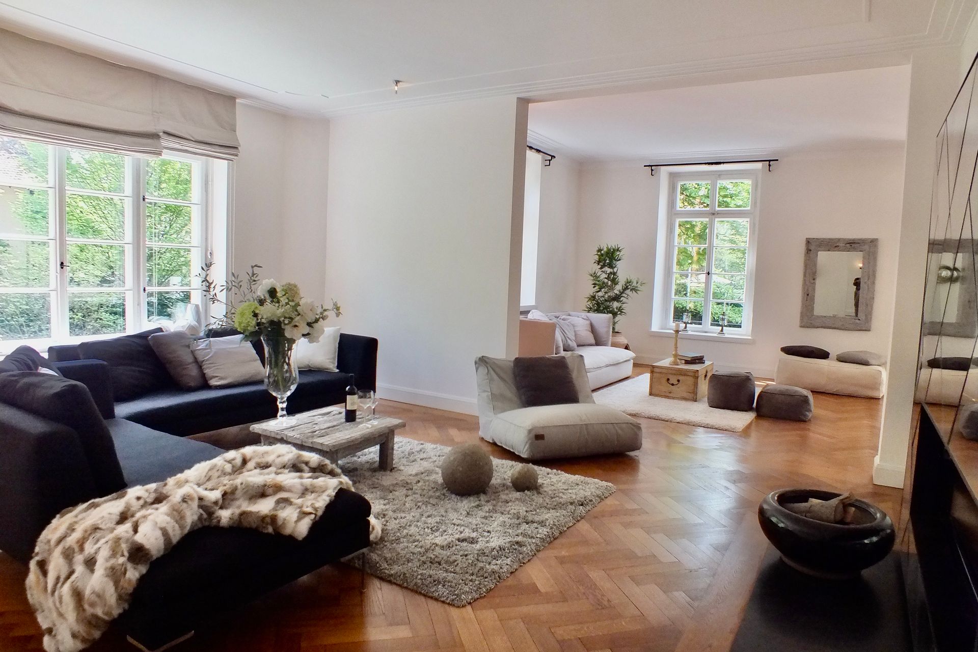 Bright living room with dark sofa, light rugs, wooden floor, and large windows with greenery outside
