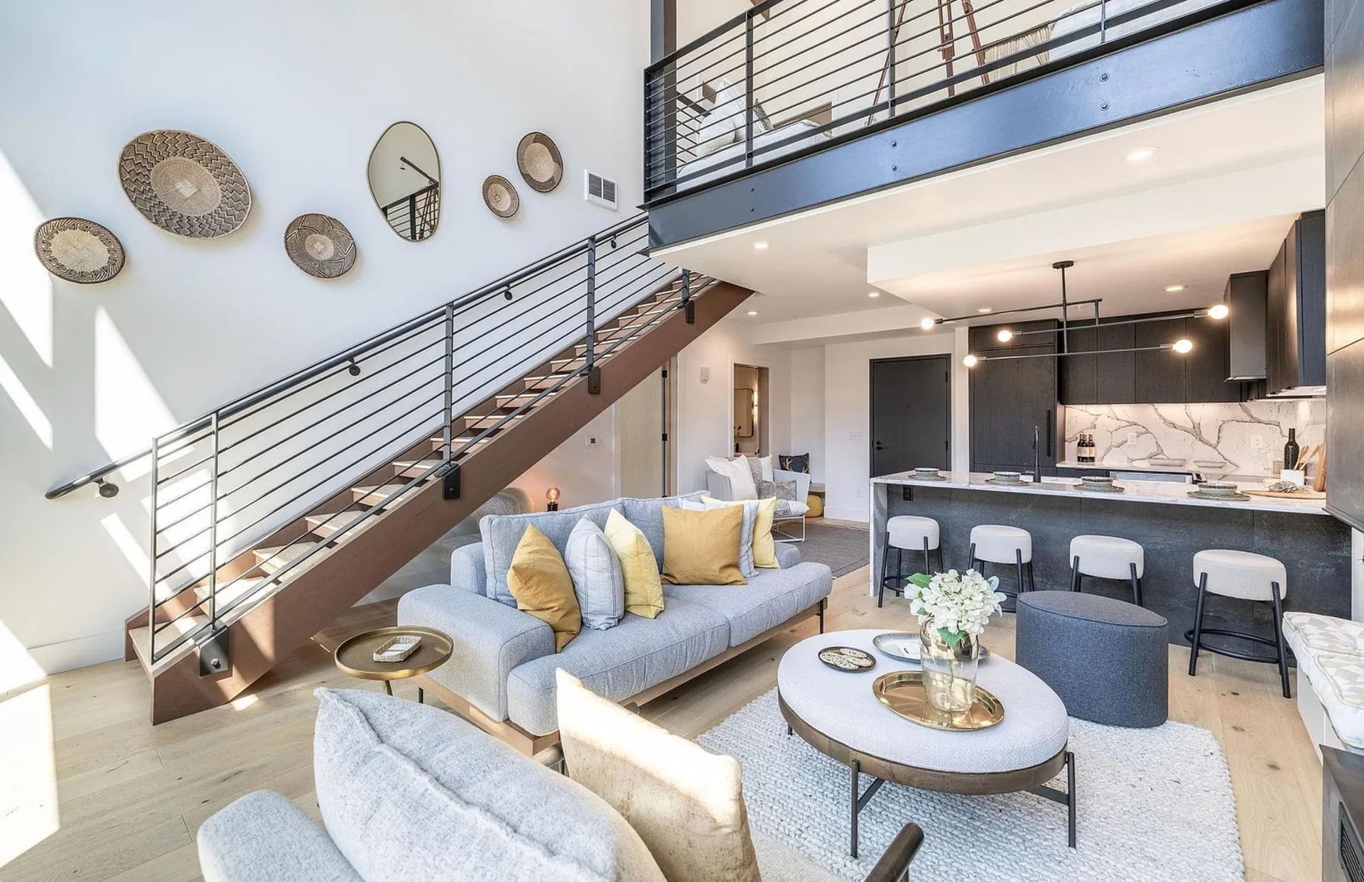 Bright modern living room with staircase, gray sofa, round coffee table, and open kitchen.