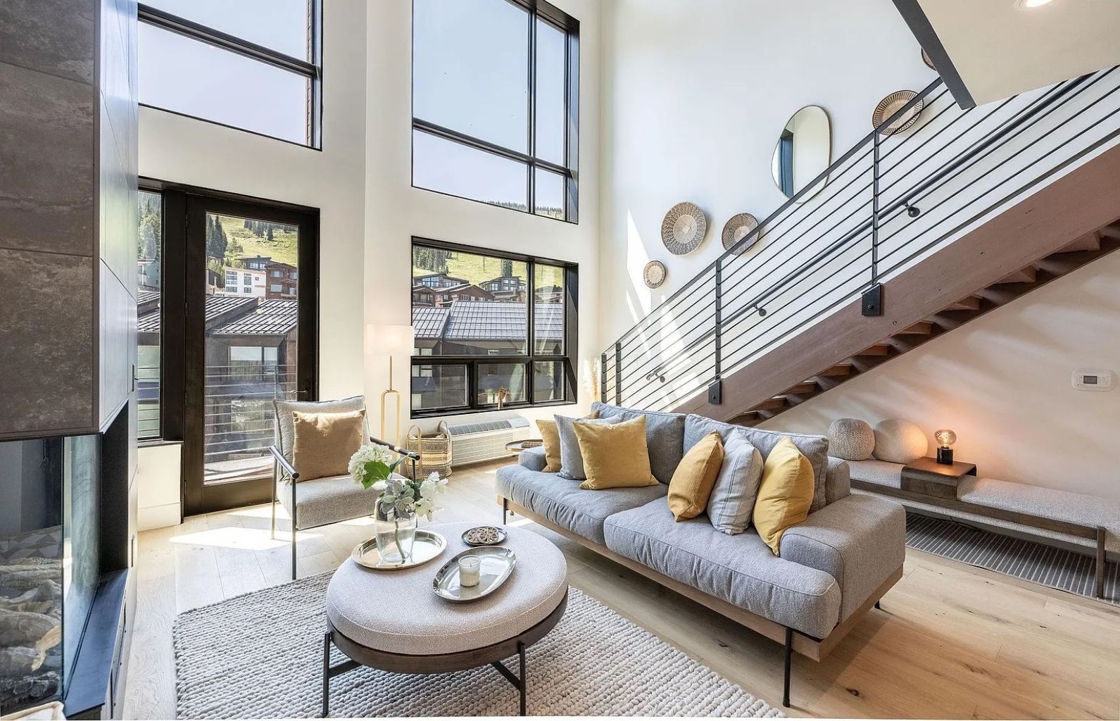 Bright modern living room with tall windows, gray sofa, round coffee table, and staircase railing above