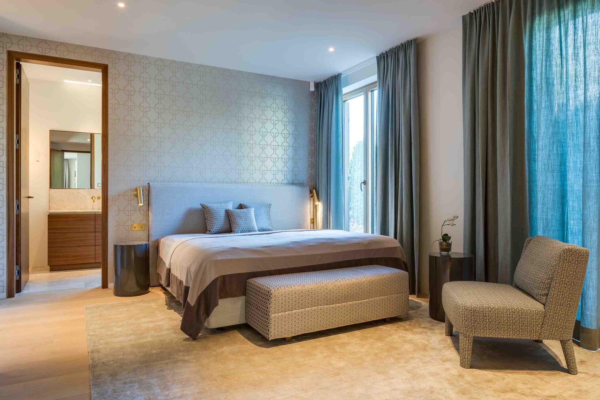 Modern bedroom with a bed, bench, armchair, large windows, and an en suite bathroom doorway.