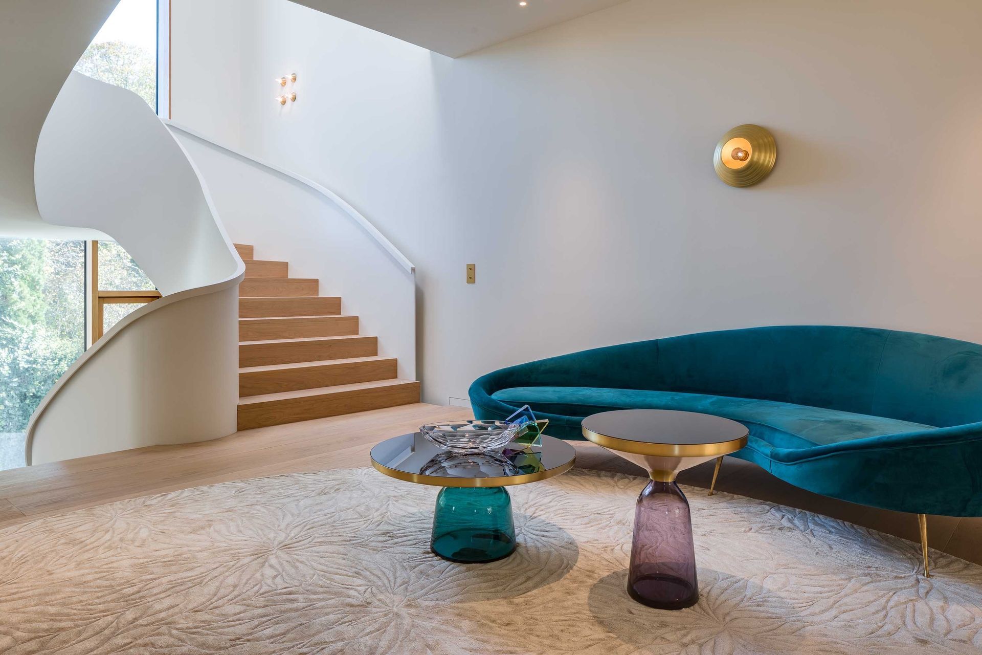 Modern living room with a curved blue sofa, spiral staircase, and glass coffee tables on a shag rug