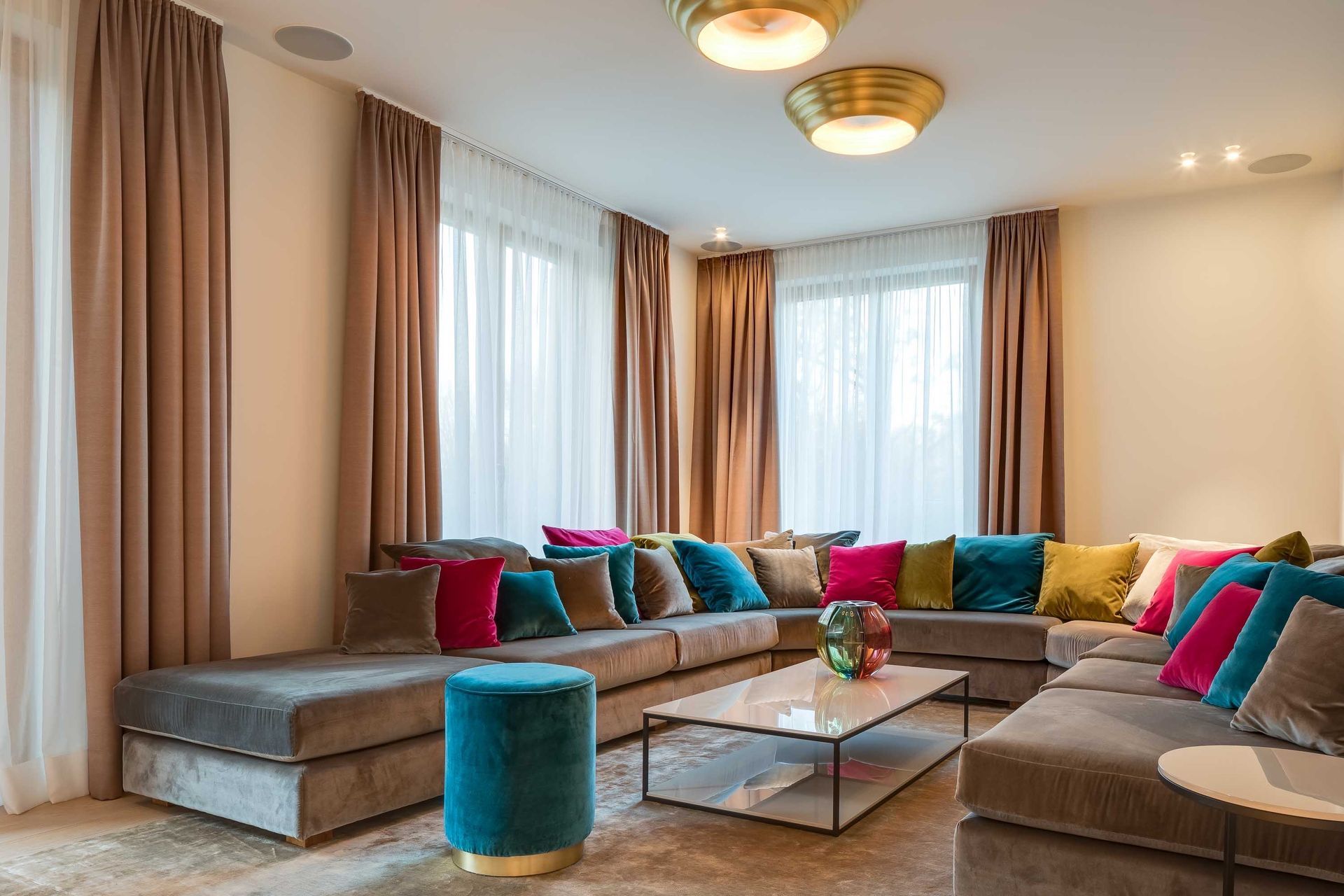 Modern living room with beige sectional sofas, teal and pink cushions, glass coffee table, and large windows