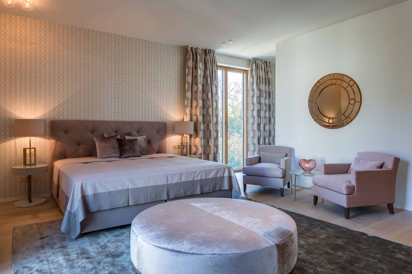 Soft beige bedroom with a neatly made bed, two chairs by the window, and a round ottoman on a rug.