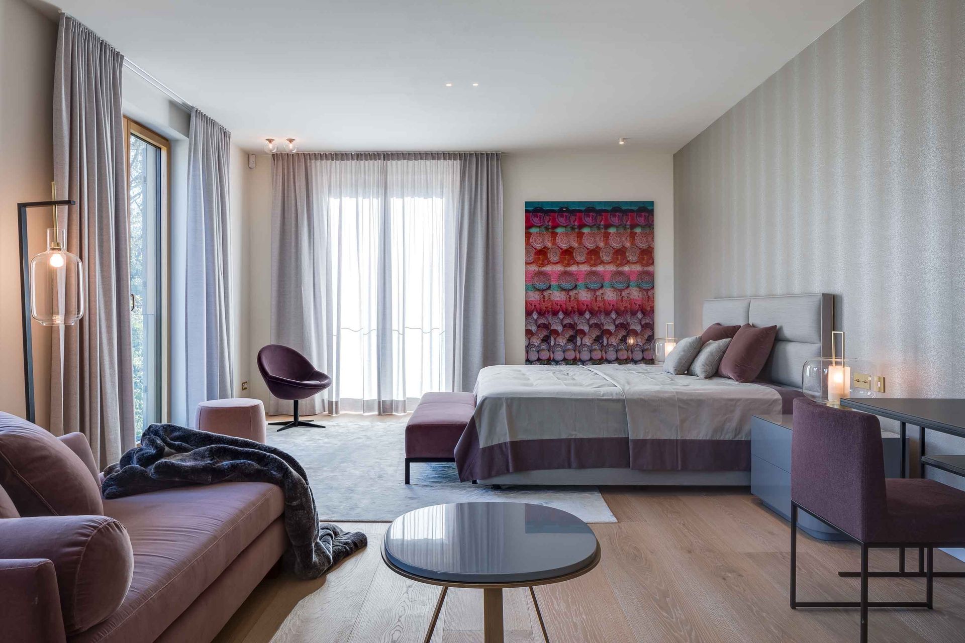 Modern hotel room with bed, sofa, chairs, and floor-to-ceiling windows with sheer curtains.