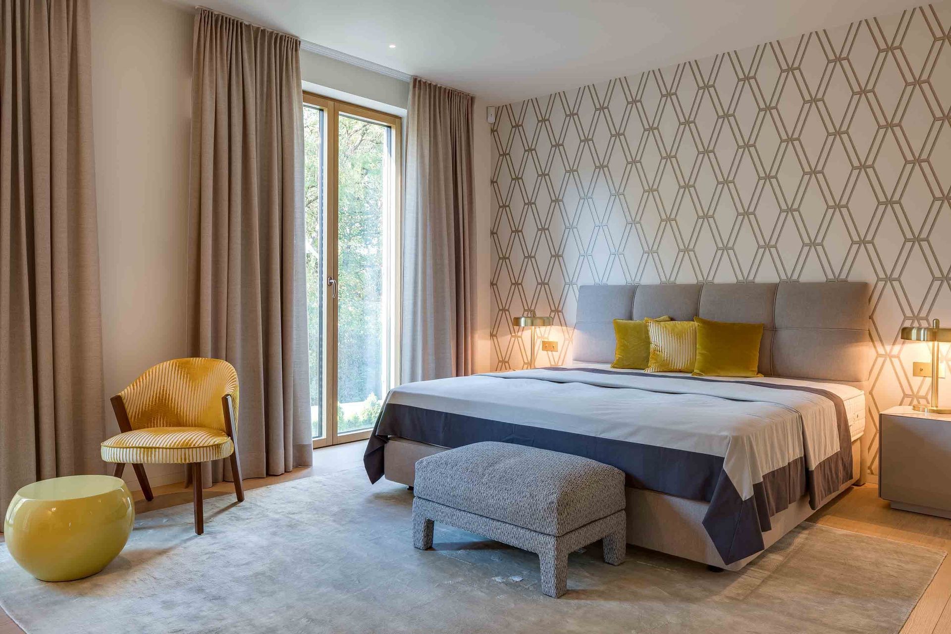 Bright bedroom with a large bed, patterned wallpaper, beige curtains, and a window chair with a yellow pillow