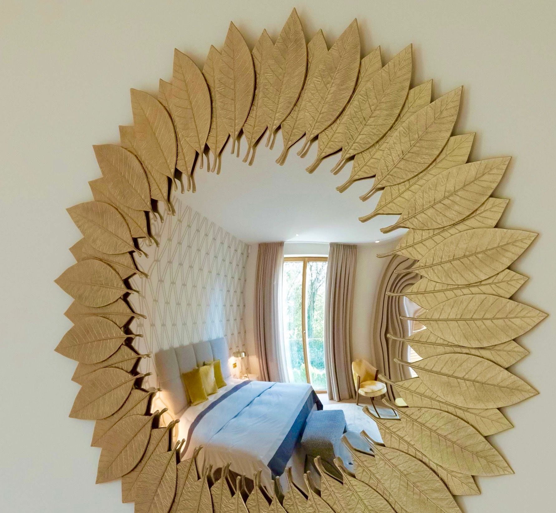 Sunburst gold mirror reflecting a bright bedroom with a bed and window curtains