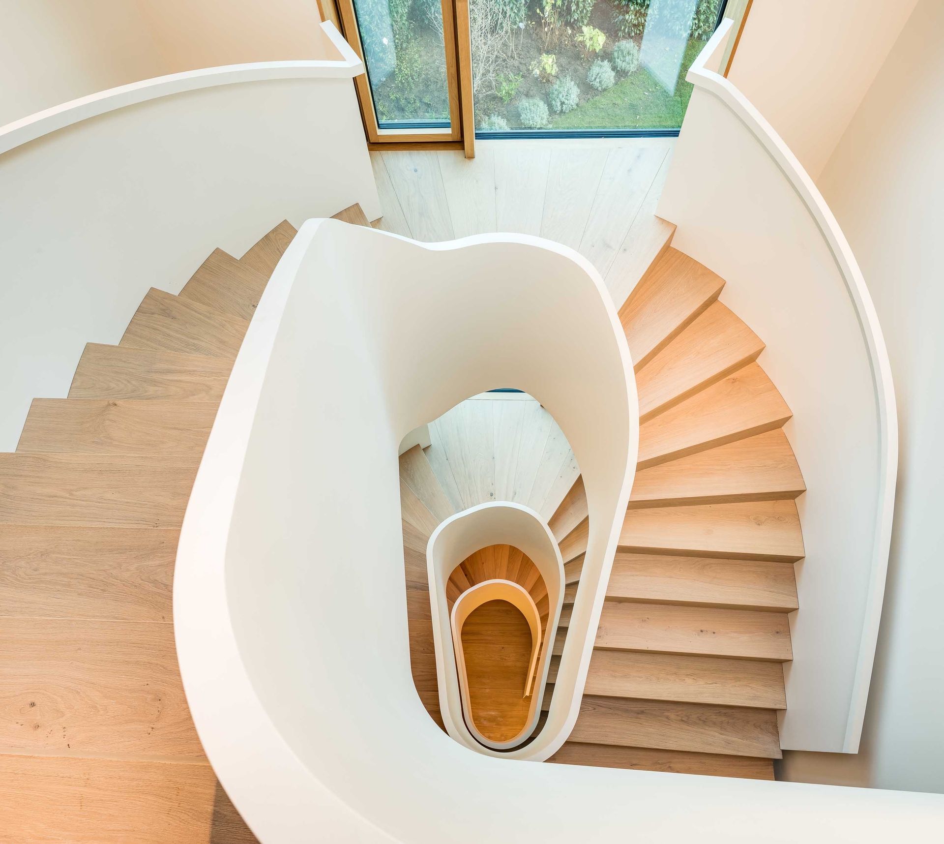 Top-down view of a curved white staircase with beige steps and a central opening, lit by a nearby window.