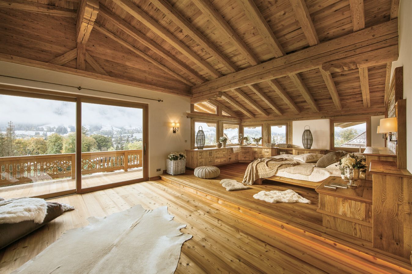 Rustic wooden bedroom with vaulted ceiling, large windows, and sunlight over a bed and cowhide rug