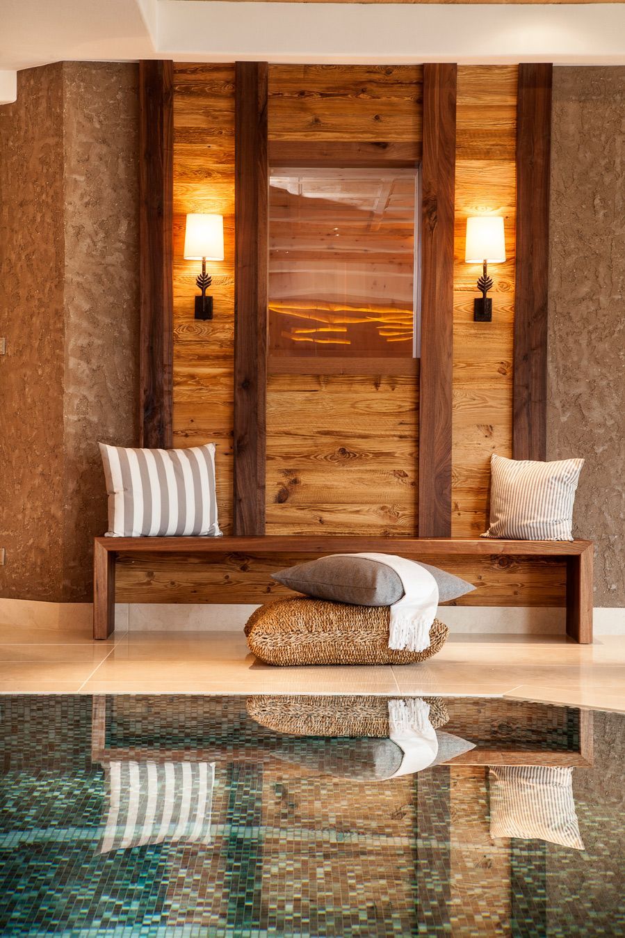 Modern spa lounge with wooden bench, striped cushions, and reflected indoor pool water on a warm stone wall backdrop