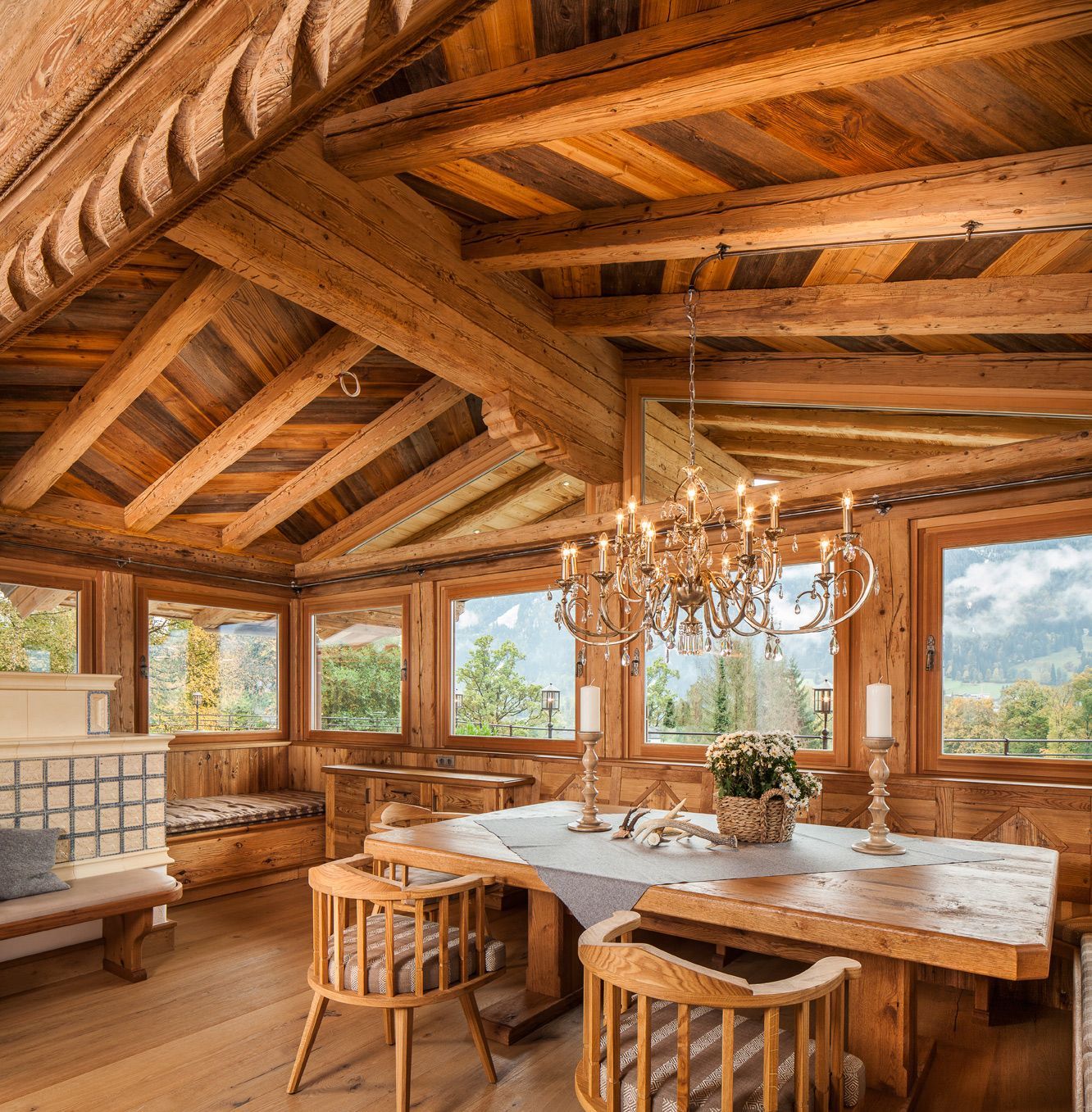 Rustic dining room with exposed wooden beams, large windows, chandelier, and a long table set with flowers.