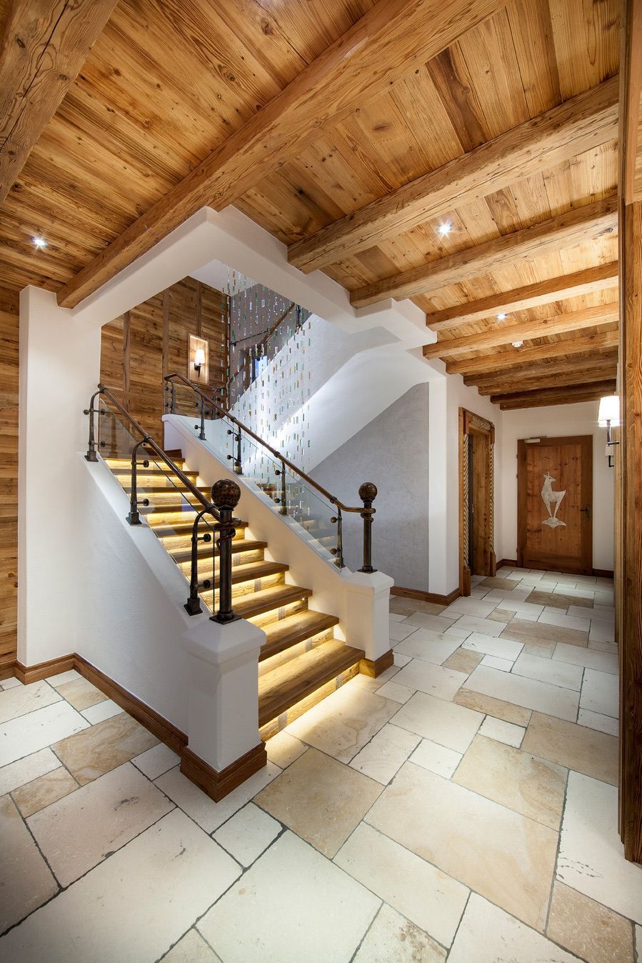 Elegant entry hall with wood ceiling, stone tile floor, and a curved staircase with lit steps