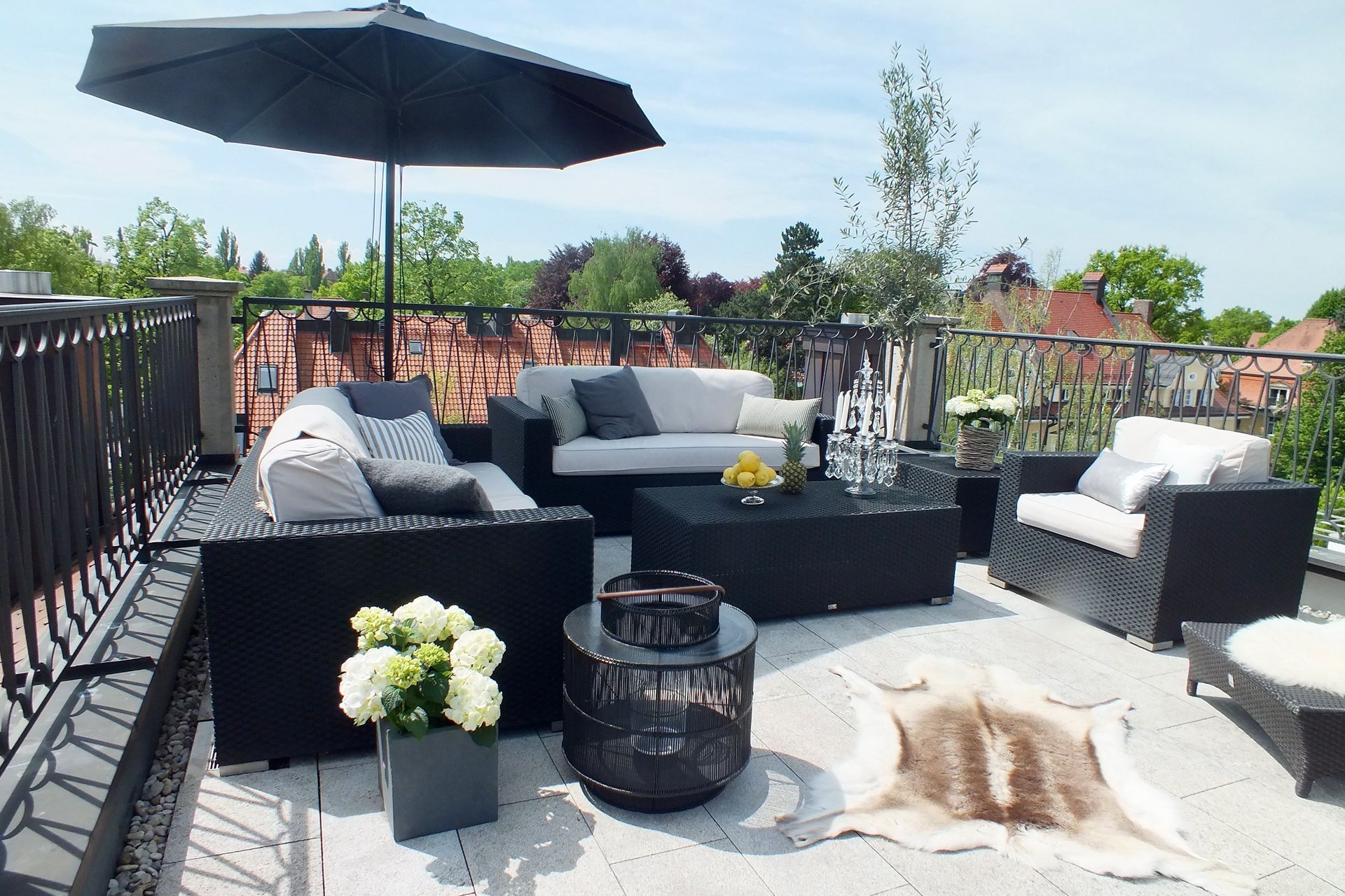 Patio seating area with black wicker furniture, umbrella, white cushions, and potted flowers under a bright sky