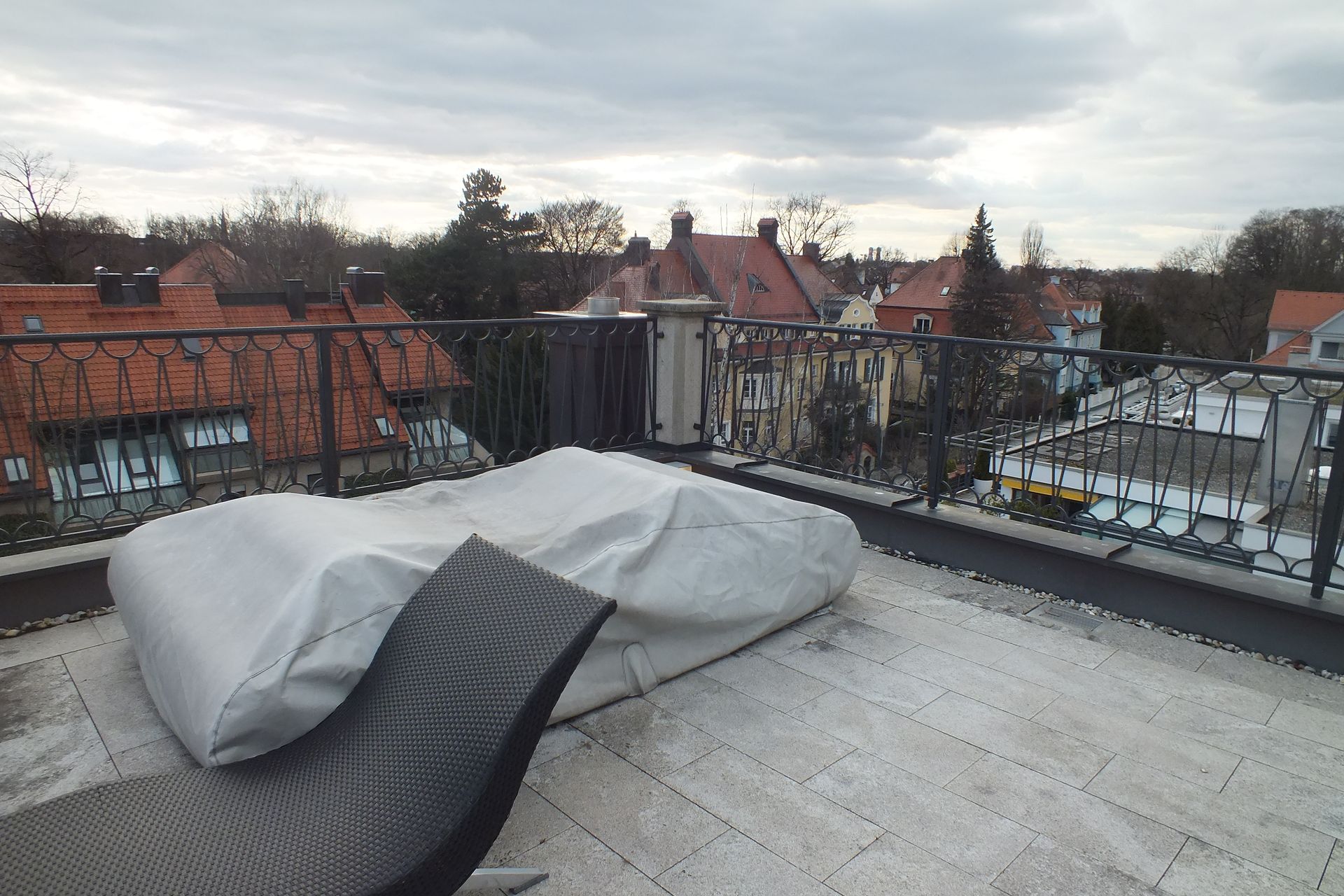 Rooftop terrace with covered lounge chairs overlooking red-tiled buildings and trees under a cloudy sky
