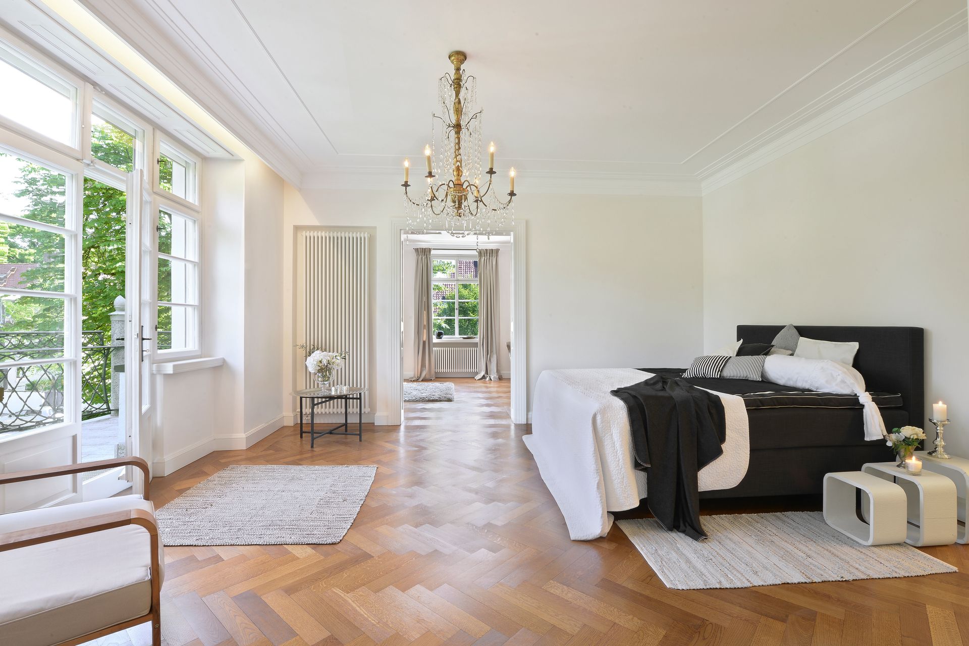 Bright bedroom with large windows, chandelier, white walls, and a black bed on a hardwood floor.