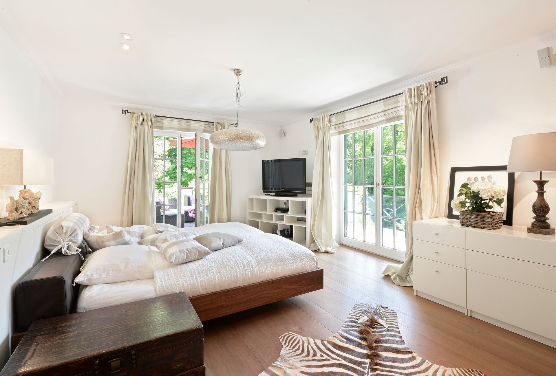 Bright bedroom with a large bed, TV, white dresser, and sunlight through tall windows and curtains