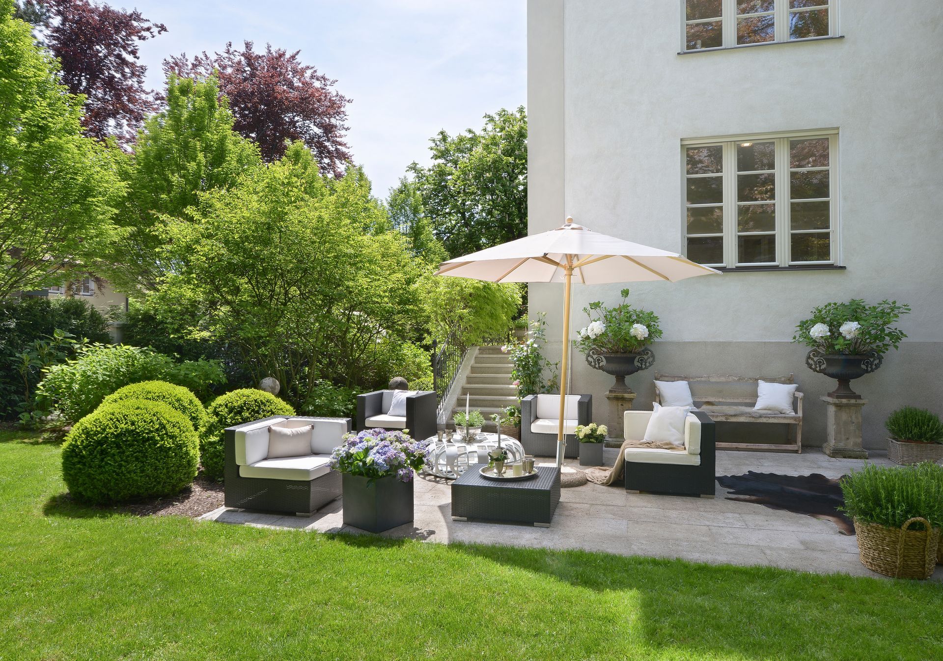Stylish patio seating with white umbrella beside a white house, surrounded by green lawn and shrubs.
